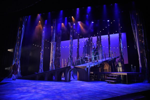 Tree trunks standing on a stage, lights strung between them, lit in purple. A wooden ramp goes along the center of the stage, along the top of which three in-costume cast members stand.