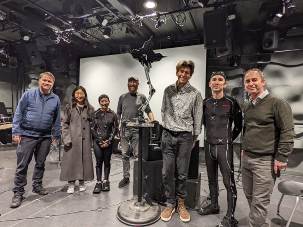 Members of the Visualizing Telematic Performance team stand on a stage after the December concert. From left to right are John Granzow, Rebecca Zhang, Sui Lin Tam, Matt Albert, Adam Schmidt, Gavin Ryan, and Michael Gurevich, with LARS the robot in the middle.