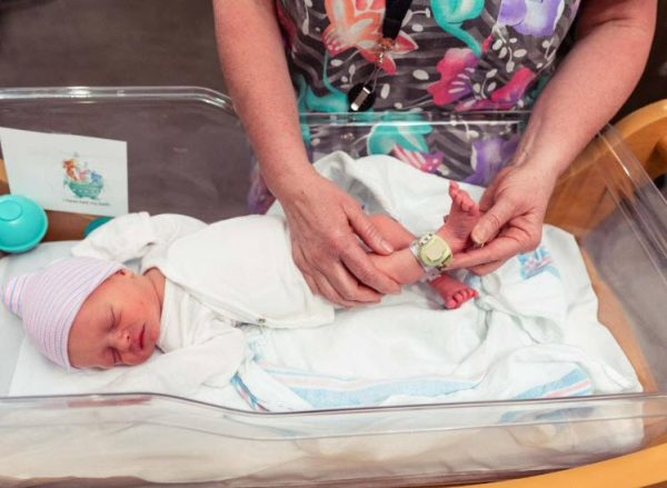 A newborn infant is shown with the HUGS tracker on their ankle. A medical professional is holding up the infants leg to show the device.