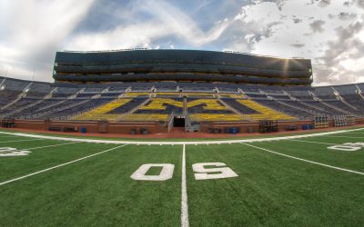 The Michigan Stadium - the perspective is on the field at the 50 yard line looking toward the bleachers and a partly sunny, partly cloudy sky.