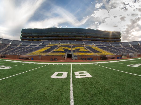 The Michigan Stadium - the perspective is on the field at the 50 yard line looking toward the bleachers and a partly sunny, partly cloudy sky.