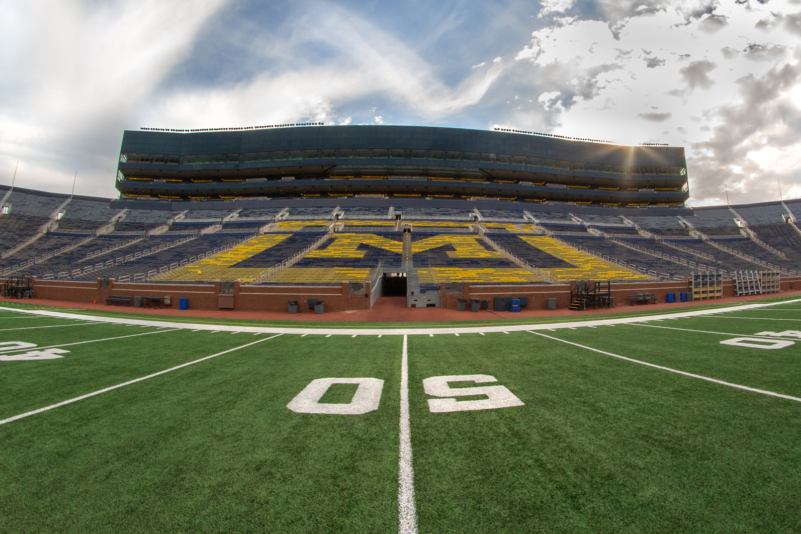 The Michigan Stadium - the perspective is on the field at the 50 yard line looking toward the bleachers and a partly sunny, partly cloudy sky.