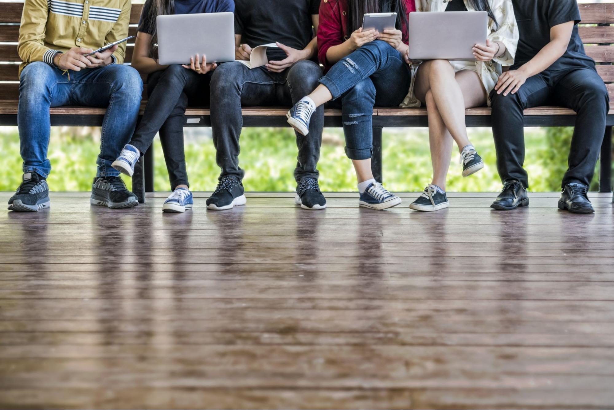 A group of students pictured from the shoulders down sitting along a wooden bench, working on their laptops.