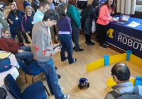 A group of people watch a small robot that is on the ground. The robot has wheels and is the size of a large grapefruit. One person has a remote control to operate the robot.