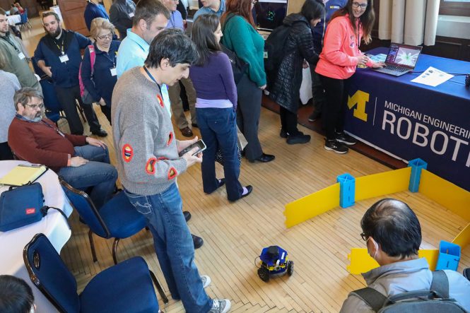 A group of people watch a small robot that is on the ground. The robot has wheels and is the size of a large grapefruit. One person has a remote control to operate the robot.