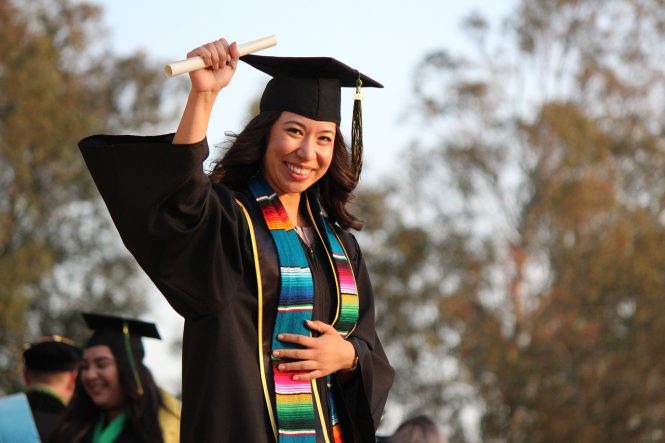 A person is wearing a black graduation robe and mortar board. They are holding their diploma victoriously in the air and they are smiling.