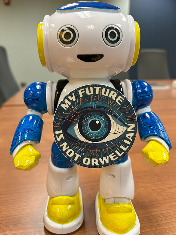 A blue, white, and yellow robot figure with round eyes and a smiling mouth has a circular sticker stuck to its chest. The sticker has a drawing of an eye in the center with the text "My Future Is Not Orwellian" wrapped around it.