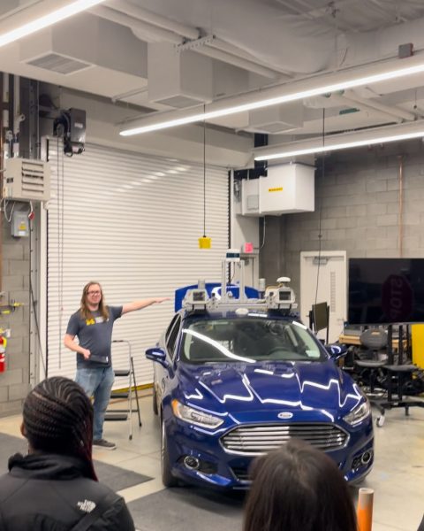 A student stands next to a blue Ford focus that is equipped with autonomous vehicle technology.
