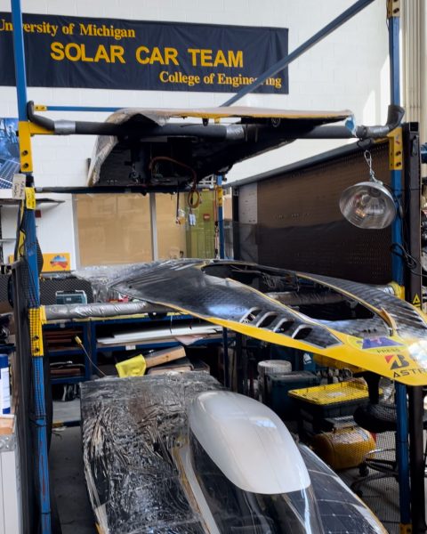 2 shells of solar cars and one completed car are stacked on a pallet rack system. The University of Michigan Solar Car team flag is maize and blue and displayed in the background.