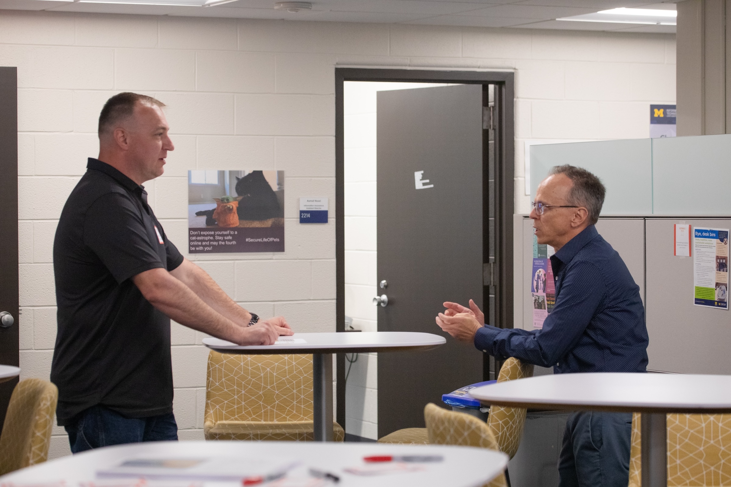 2 men have a discussion across a small round table. One is standing and one is sitting. They are both wearing navy blue shirts.