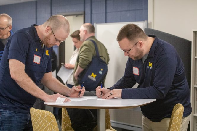 2 men wearing navy blue lean over a small round table, writing on paper.