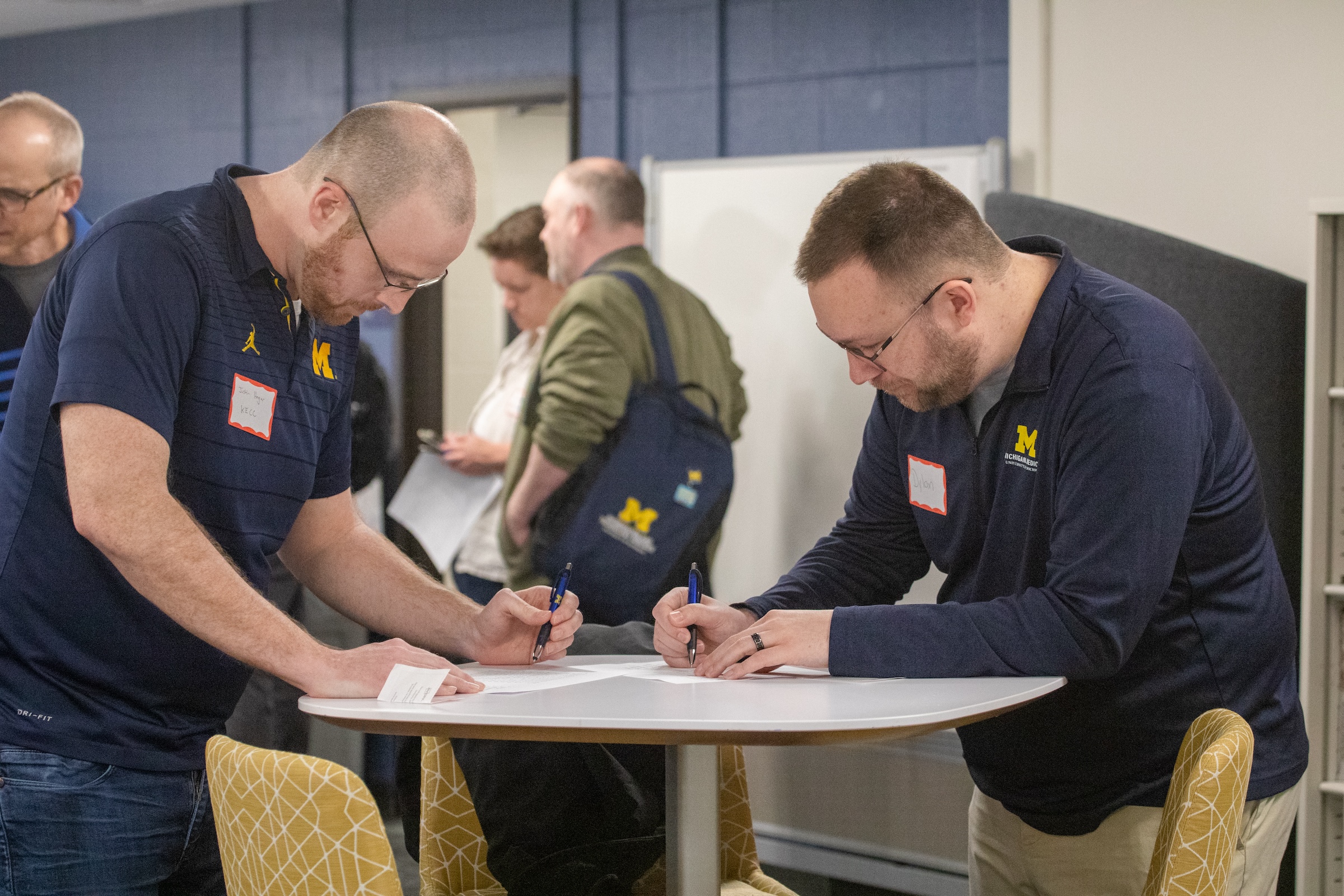 2 men wearing navy blue lean over a small round table, writing on paper.