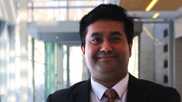 Alauddin Ahmed stands in a hallway with the photo showing from his shoulders up. He is wearing a black suit jacket, a striped shirt, and a tie. 