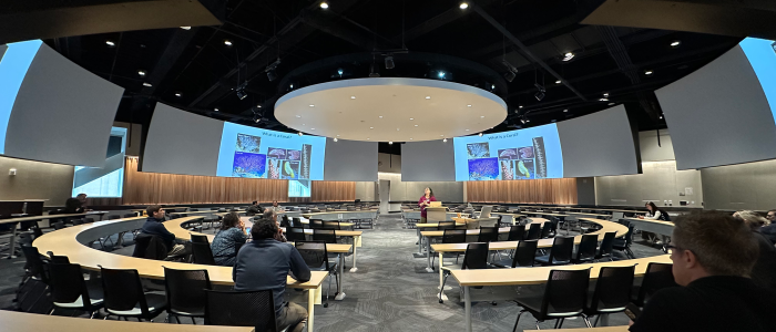 A 360-degree classroom with an instructor teaching from the center of the room. Class slides and videos are displayed on monitors that ring the classroom.