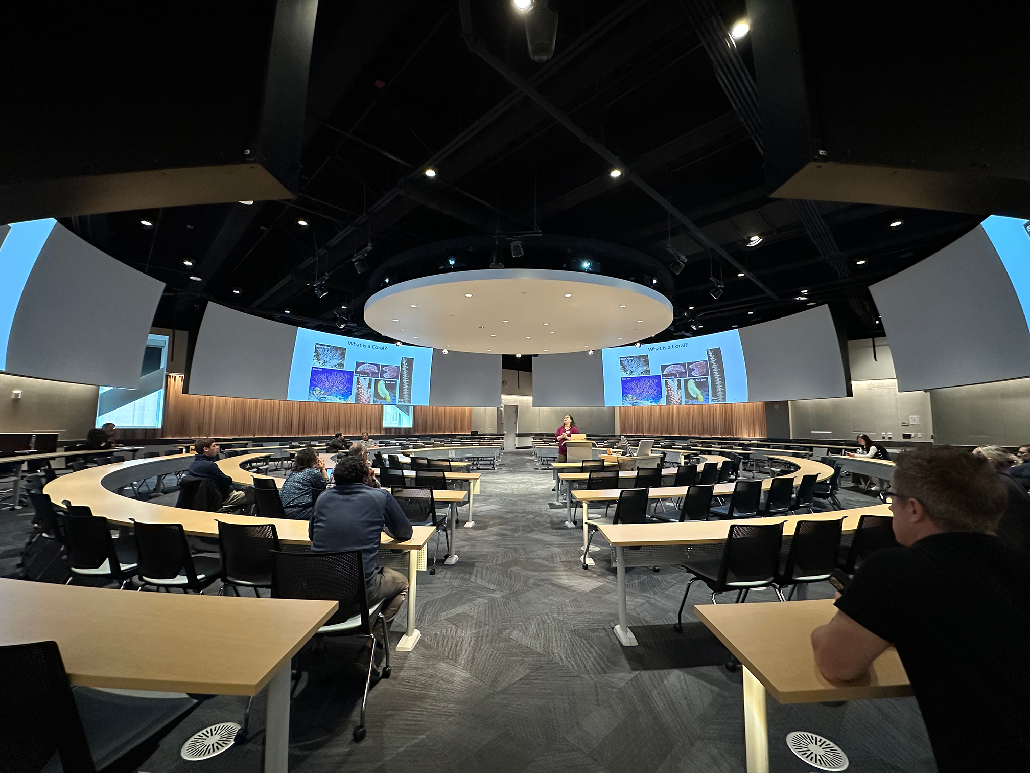 A 360-degree classroom with an instructor teaching from the center of the room. Class slides and videos are displayed on monitors that ring the classroom.