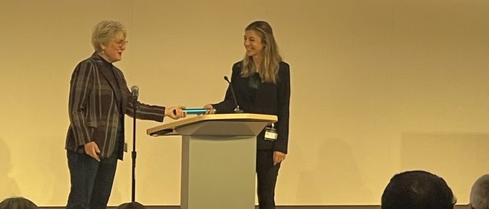 Outgoing Michigan IT Chair Kerry Flynn, CIO of Ross School of Business passes the baton (literally) to incoming Chair Aubrey Morse on stage in front of 2024 Winter Innovation Expo attendees.