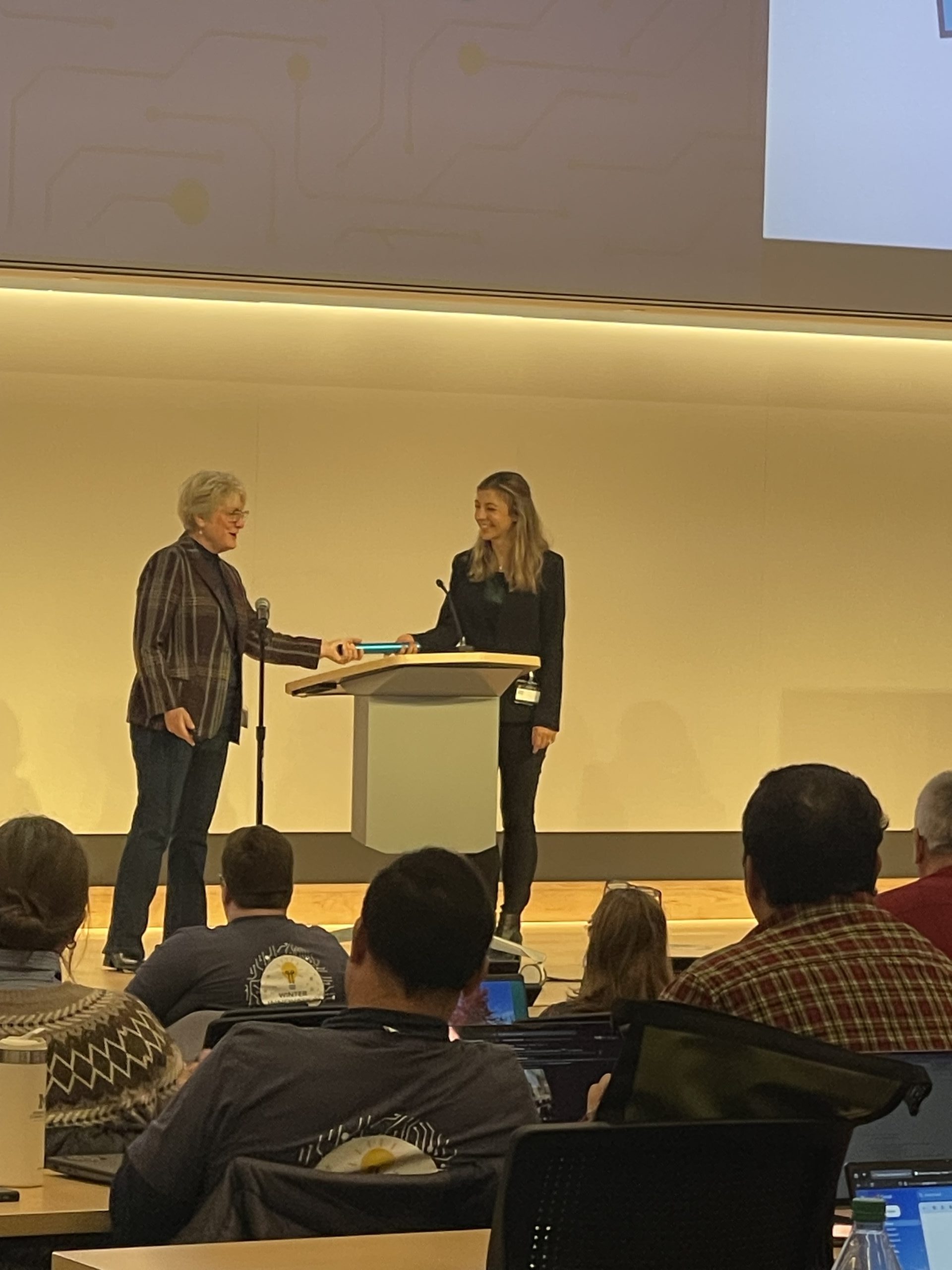 Outgoing Michigan IT Chair Kerry Flynn, CIO of Ross School of Business passes the baton (literally) to incoming Chair Aubrey Morse on stage in front of 2024 Winter Innovation Expo attendees.