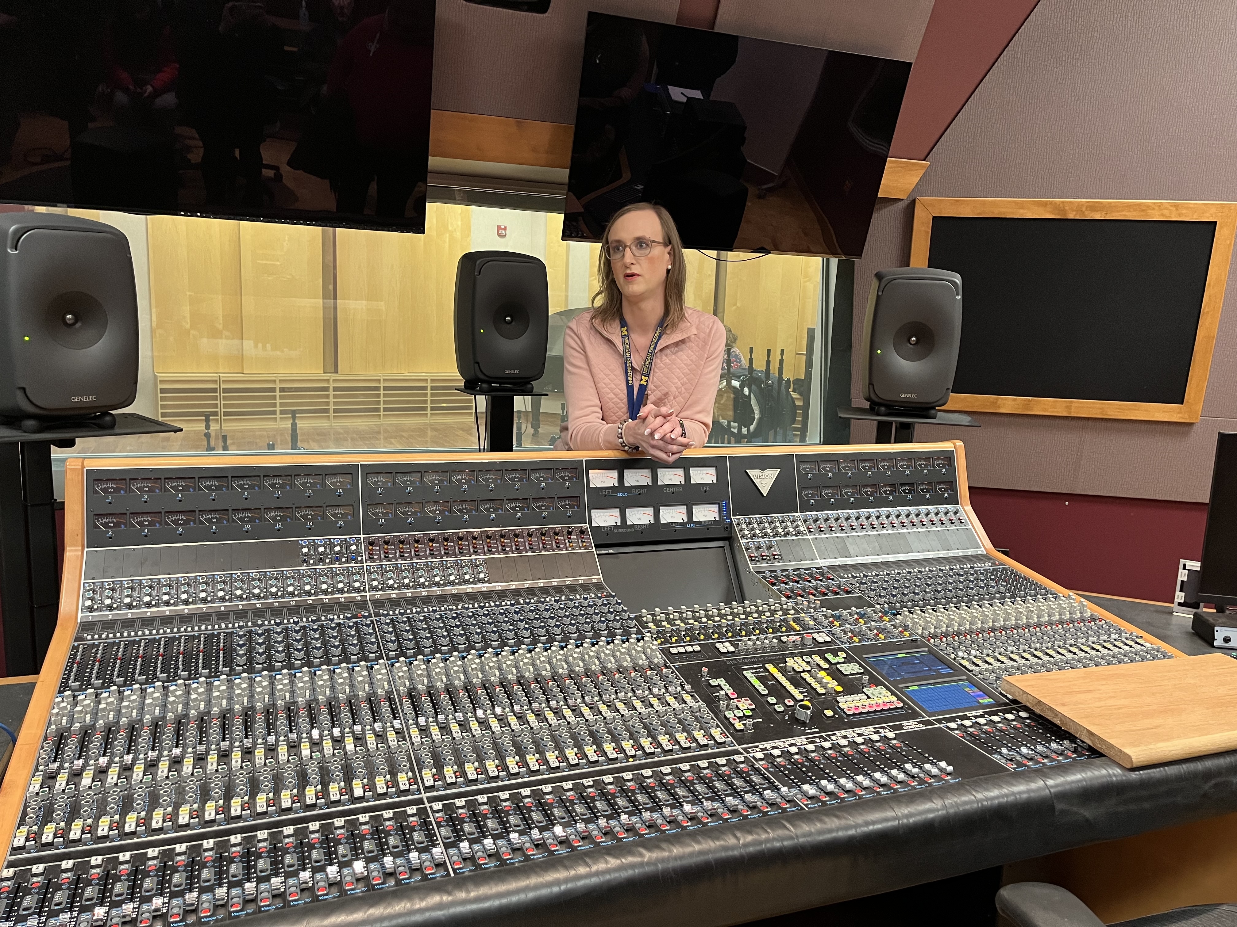 A large switchboard inside a recording studio with tv screens and speakers above it. A woman stands leaning on the edge of the board.