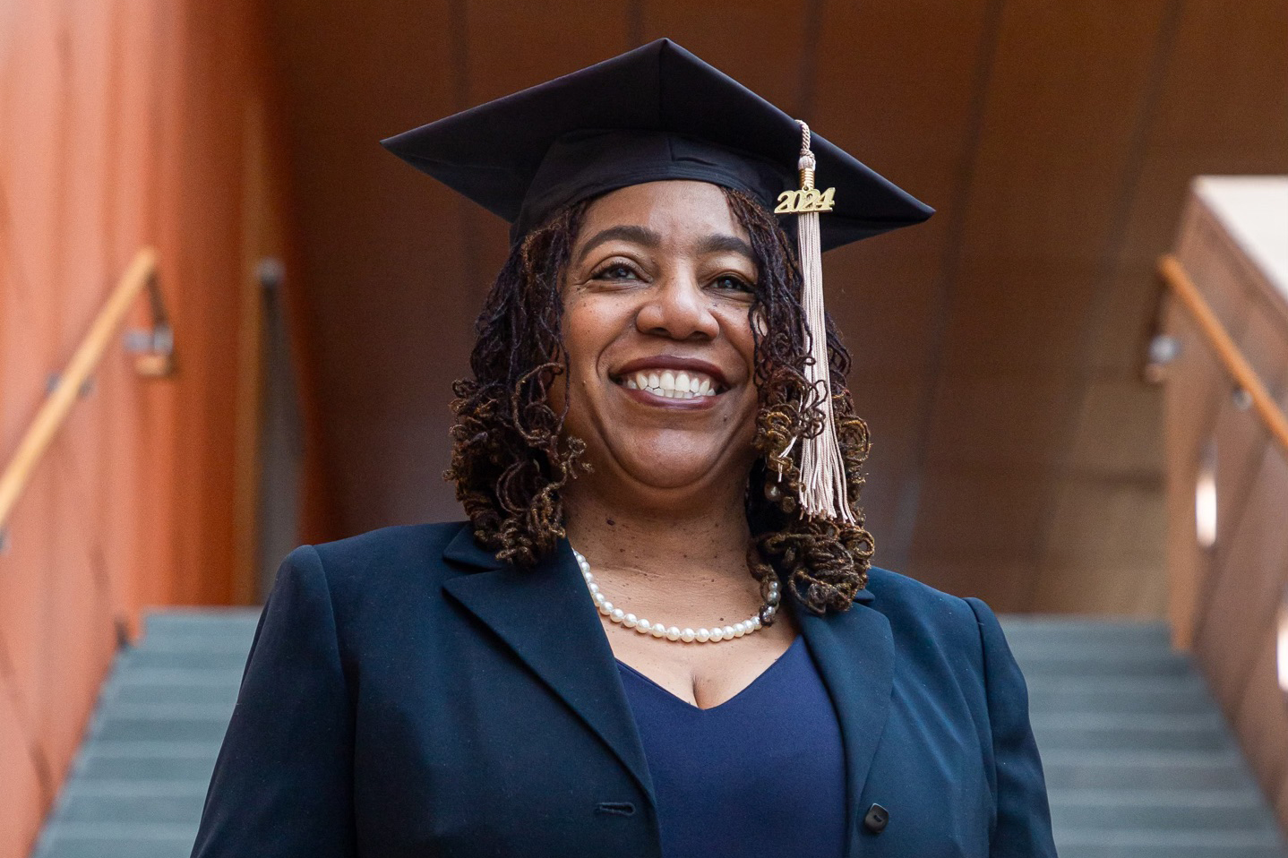 Lovida Roach pictured smiling in a blue dress and blazer, wearing a pearl necklace, and donning her Graduation cap for her MBA.