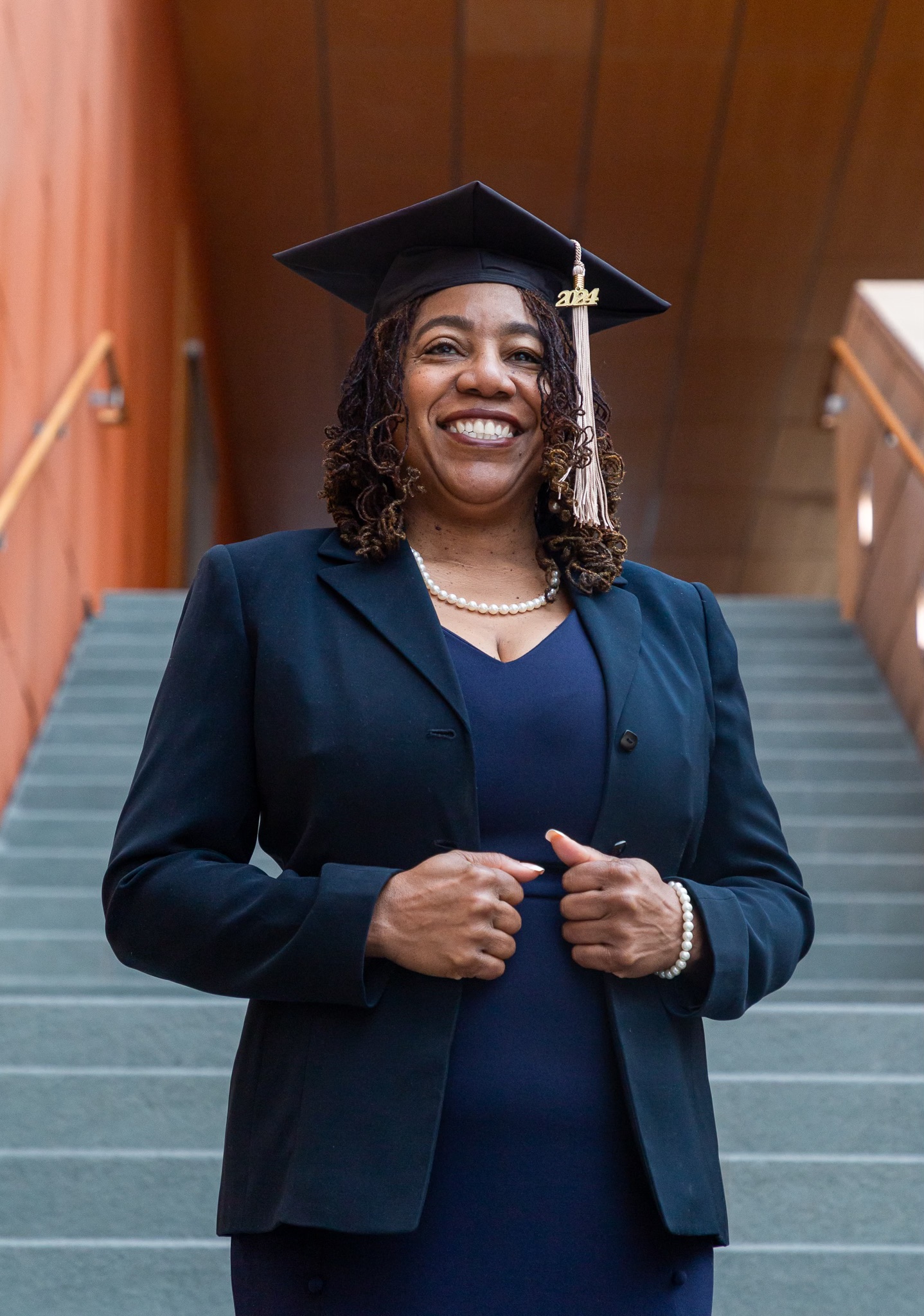 Lovida Roach pictured smiling in a blue dress and blazer, wearing a pearl necklace and bracelet, and donning her Graduation cap for her MBA.