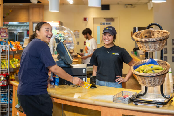 Two people stand at a counter in a dining hall, smiling, next to a basket filled with colorful fruit.