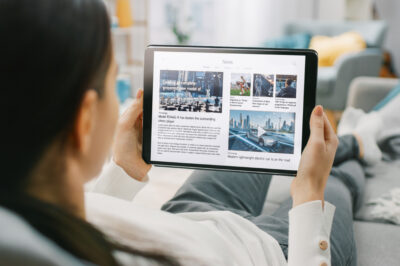 Over the Shoulder Shot of a Young Woman at Home Lying on a Couch and Using Tablet Computer in Horizontal Landscape Mode for Reading News about Technology.