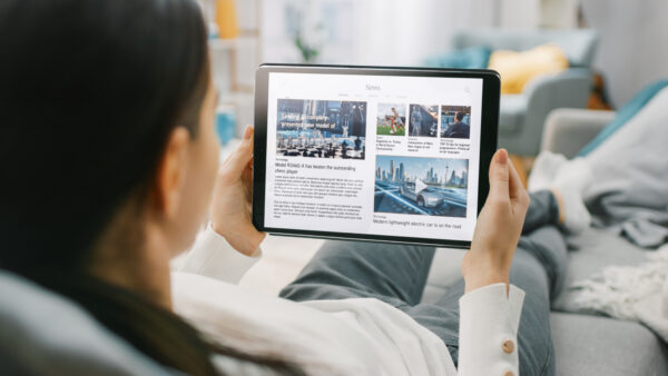 Over the Shoulder Shot of a Young Woman at Home Lying on a Couch and Using Tablet Computer in Horizontal Landscape Mode for Reading News about Technology.