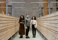 Three students browsing a card catalog aisle in a modern library setting.