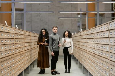 Three students browsing a card catalog aisle in a modern library setting.