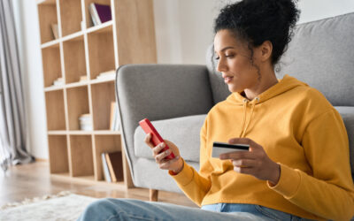A woman is seated at a desk with her phone in one hand and a credit card in the other hand.