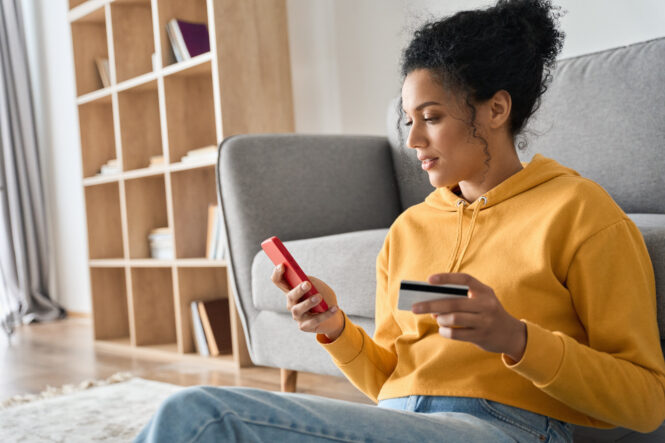 A woman is seated at a desk with her phone in one hand and a credit card in the other hand.