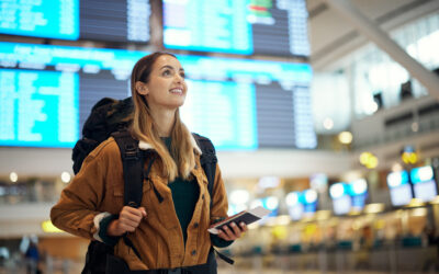 A woman carrying a backpack stands in an airport terminal with her phone, smiling.