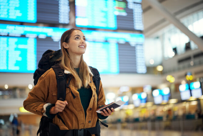 A woman carrying a backpack stands in an airport terminal with her phone, smiling.