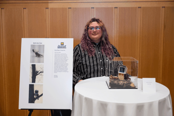 Kade Kuba standing with her "Bird's Eye View" mixed media sculpture that depicts a robin with the head of an old-timey security camera.