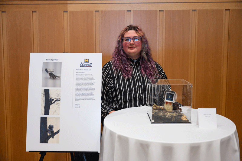 Kade Kuba standing with her "Bird's Eye View" mixed media sculpture that depicts a robin with the head of an old-timey security camera.