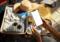 A person holds a cellphone over an open suitcase filled with clothing items for a trip.