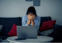 A distressed woman sits on a couch with her head in her hands. There is an open laptop on the coffee table in front of her.