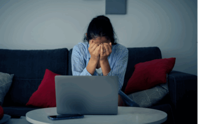 A distressed woman sits on a couch with her head in her hands. There is an open laptop on the coffee table in front of her.