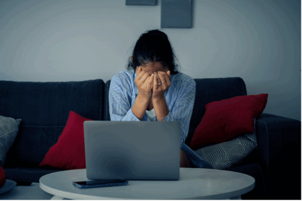 A distressed woman sits on a couch with her head in her hands. There is an open laptop on the coffee table in front of her.