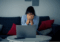 A distressed woman sits on a couch with her head in her hands. There is an open laptop on the coffee table in front of her.