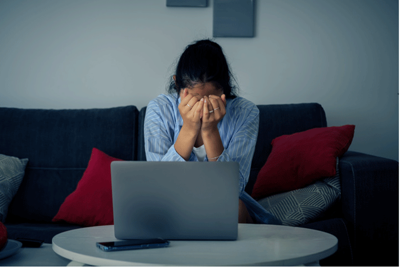 A distressed woman sits on a couch with her head in her hands. There is an open laptop on the coffee table in front of her.